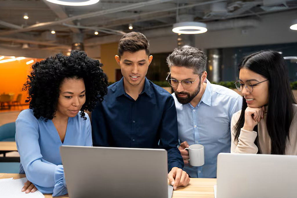 Four diverse tech professionals (a diverse team of international colleagues) smiling and reviewing data analytics on laptop and computer monitors in a bright, modern cool office workspace